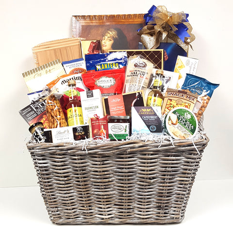 A large bold wicker hamper gift basket is overflowing with a cheese board, cheeses, gourmet sauces, Gazzosa sparkling water, chocolates, Hungarian Salami, nuts, crackers, cookies, Sea Change Smoked Salmon, mustards and lots more.