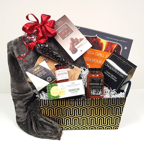 A warm cuddly blanket is nestled in a beautifully designed gourmet gift basket of black and gold along with a pizza stone, gourmet pizza sauce, crackers, and cheese, a mini cheese board, savoury shortbread, red pepper jelly and truffles and chocolates.